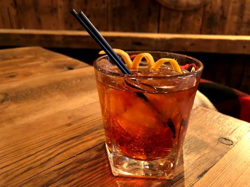 Image: A Negroni cocktail served in a rocks glass is garnished with an orange peel twist. There are two black cocktail straws in the glass. The glass sits on a wood table, behind which is a wood wall. Photo by Remington Rock. 