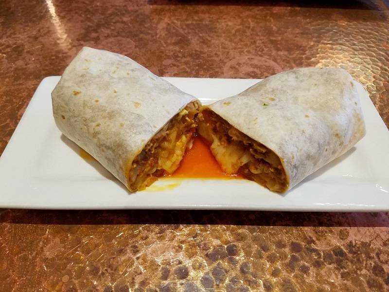 Image: A breakfast burrito is cut in half and sits on a rectagular white plate. The flour tortilla is white, and from inside the burrito is melty cheese, rice, and eggs. Everything spilling out is an orange color, and orange grease and liquid is pooled in between the burrito halves. The plate sits on a hammered copper table. Photo by Matthew Macomber. 