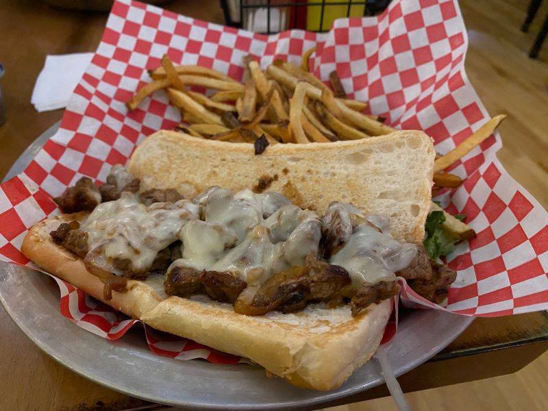 Image: An open-faced sandwich on a rectangular hoagie roll. The roll is covered in pieces of steak and topped with melted cheese. The remainder of the plate is covered in thinly cut french fries. The plate is metal with a red and white checked paper liner. Photo by Julie McClure.