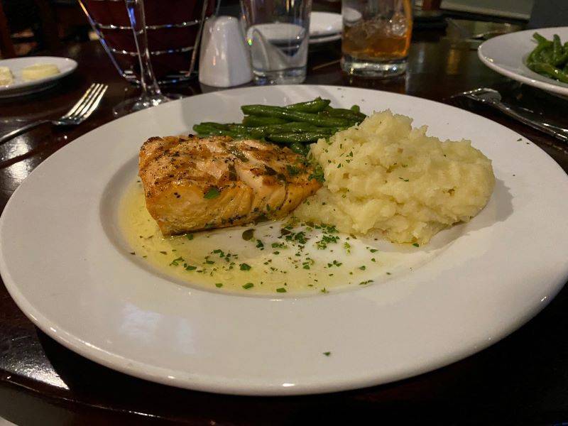 Image: A round white plate sits on a dark wood table. There is a piece of salmon, surrounded by mashed potatoes and green beans. There is lemon butter sauce on the plate, sprinkled with parsley. Photo by Julie McClure.