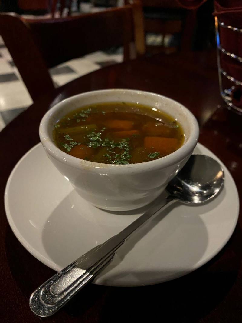 Image: A small white bowl sits on a white saucer on a dark wood table. It is filled with brown broth, and there are pieces of carrot and beef. Parsley is sprinkled on the surface. There is a metal spoon sitting on the saucer. Photo by Julie McClure.