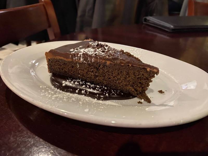 Image: An oval white plate sits on a dark wood table. There is a wedge of chocolate torte with a layer of chocolate ganache. It sits on a base of chocolate sauce, and is sprinkled with powdered sugar. Photo by Julie McClure.