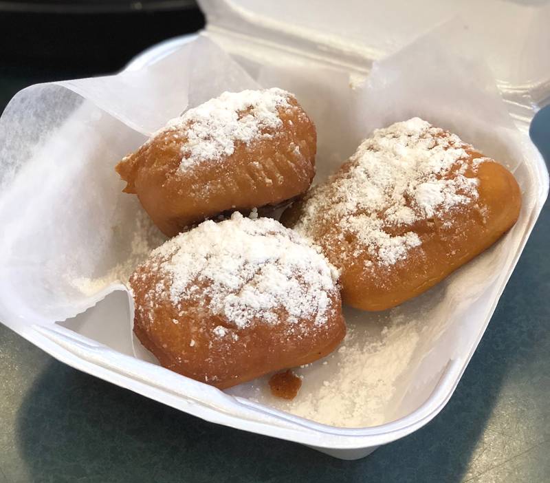 Image: Three beignets from Stango Cuisine sit in a square, white styrofoam container. The beignets are golden colored and generously dusted with powdered sugar. Photo by Jessica Hammie. 