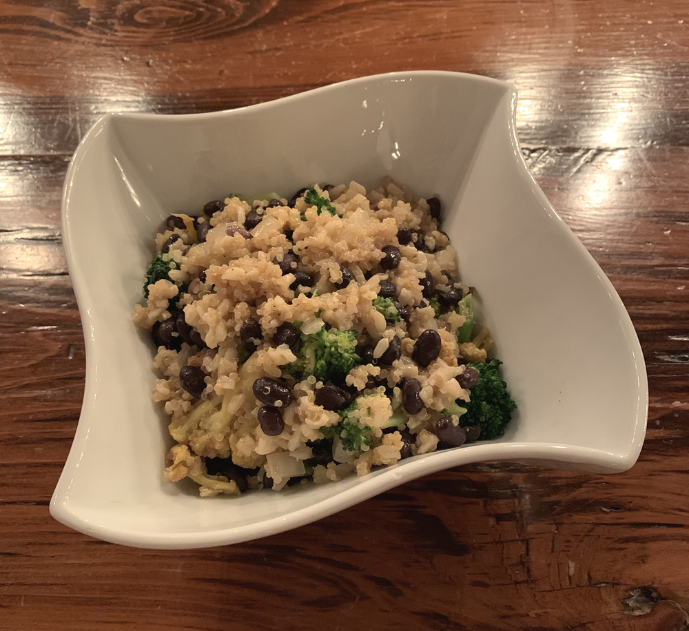 Image: Curry cauliflower and brown rice bowl at the WheelHouse. The bowl is white and a wavy square shape. In the bowl are brown rice, quinoa, broccoli, black beans, and cauliflower. The bowl sits on a wood table. Photo by Jessica Hammie.