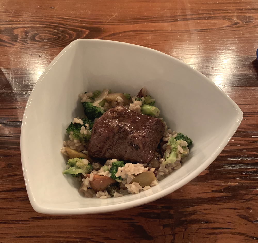 Image: Braised beef short rib with brown rice and vegetables at the WheelHouse. In a white, rounded triangular bowl are brown rice, broccoli, carrots, and a square piece of beef short rib. The bowl sits on a wood table. Photo by Jessica Hammie.