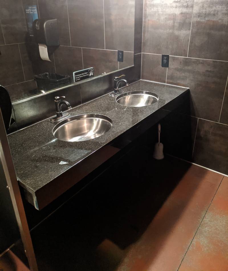 Image: A double sink with black marble counter and stainless steel sinks. A large rectangular mirror is above the sink. The floor is brown and the wall is large grayish brown tiles.