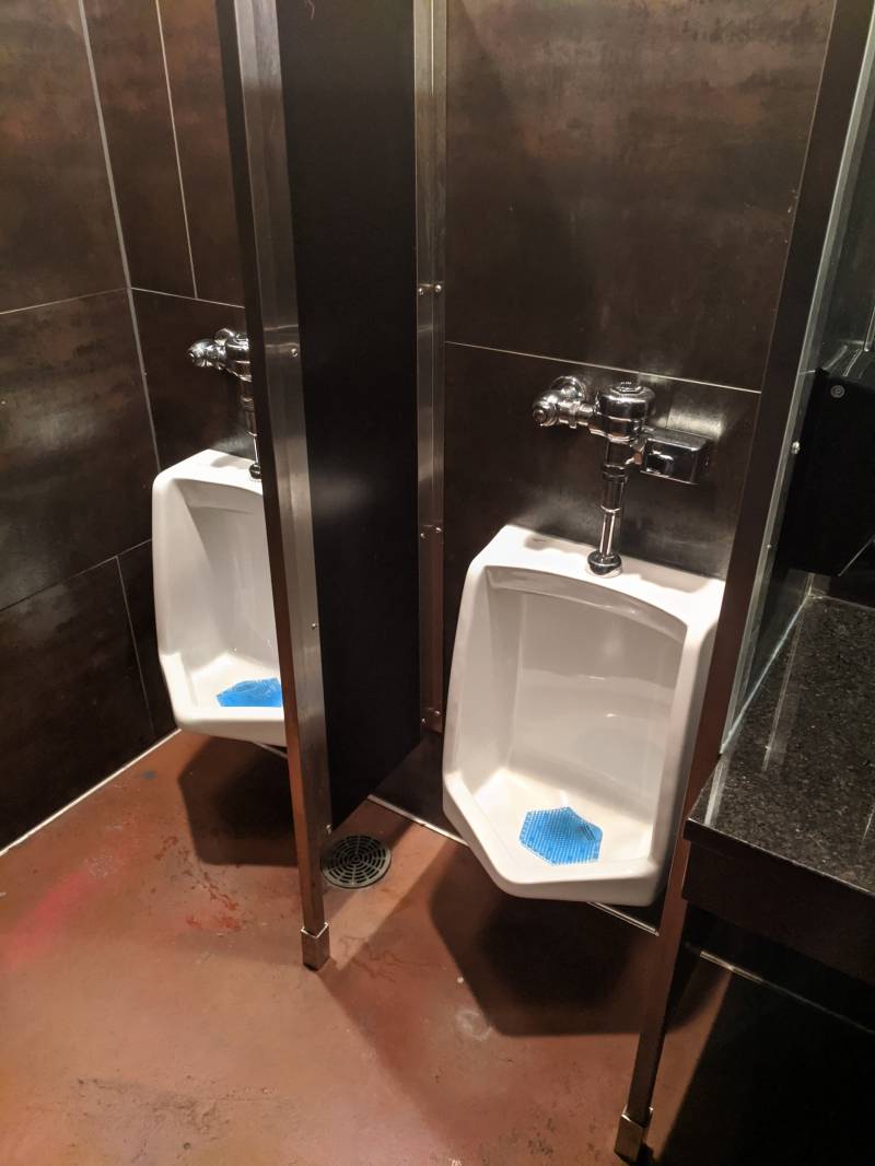 Image: Two white porcelain urinals separated by narrow black wall. The floor is brown and the wall has large grayish brown tiles.