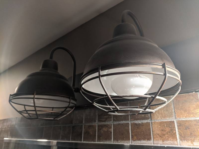 Image: Two lights are attached to a wall. The lights have round metal cage-like covers. The top portion of the wall is painted dark gray. Below the painted portion are two rows of small light brown square tiles.