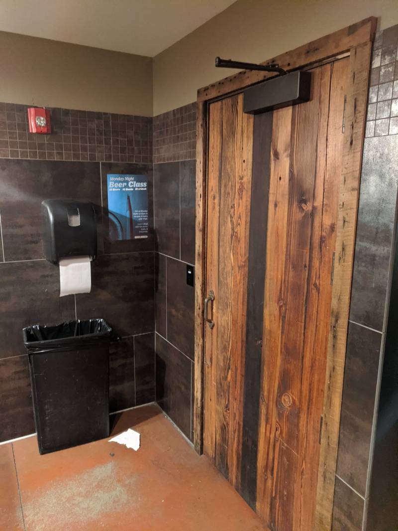 Image: The corner of a bathroom. There is a large wooden door to the right. To the left is a black rectangular garbage can with a black paper towel dispenser above it. The walls have dark grayish brown tile.