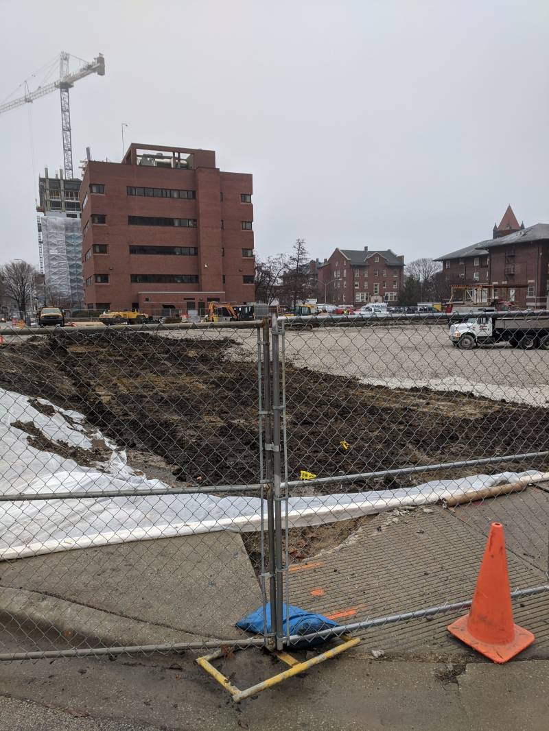 Image: A construction area surrounded by a chain link fence. There is an orange safety cone in the foreground. Behind the fence is a large hole in the ground. Beyond the hole you can see buildings and a crane.