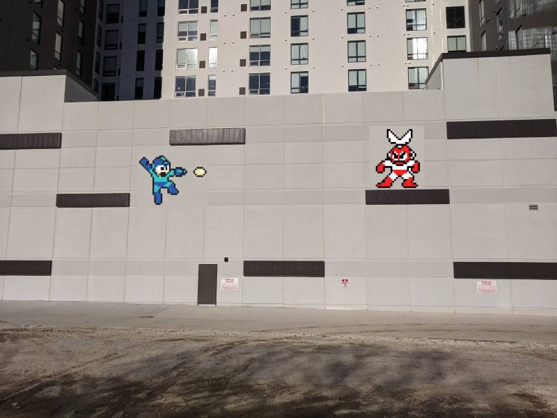 Image: A mostly light gray wall with several dark gray horizontal rectangles placed sporadically. The writer added images of a blue Mega Man character and red and white Mega Man character. An empty parking lot is in the foreground.