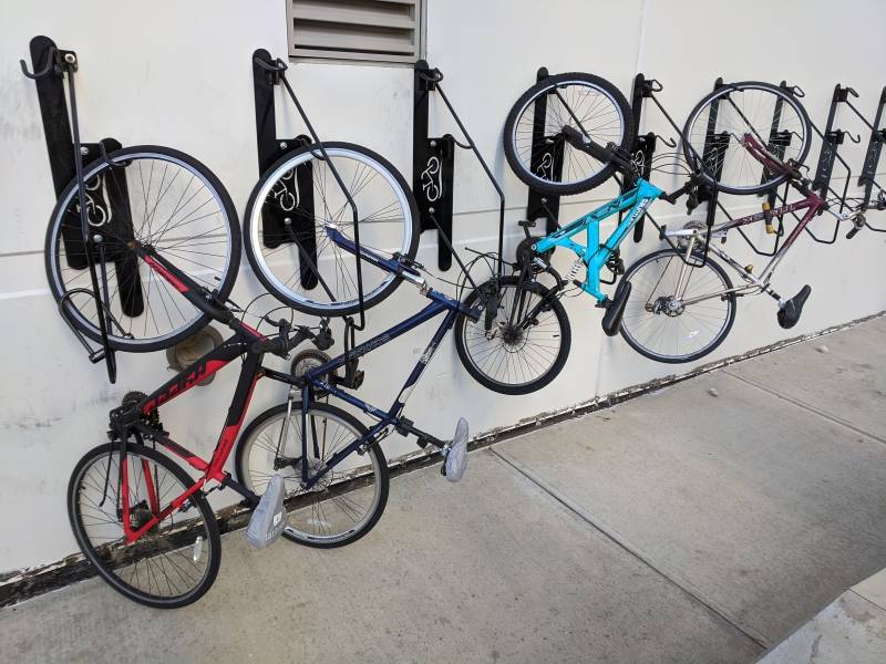 Image: Several black bike hooks are attached to a white wall. There are four bikes, each hanging from a hook by the front wheel.