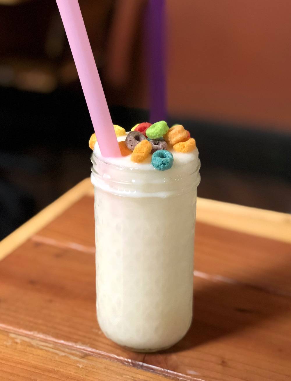 Image: Watson's Toucan Slam boozy slushie sits on a wood table. It is a frozen, blended white beverage served in a mason jar and garnished with Froot Loopsâ„¢ cereal pieces. A pink straw and a purple straw stick out of the drink on opposite sides of the glass. The glass is on a wood table. Photo by Jessica Hammie.