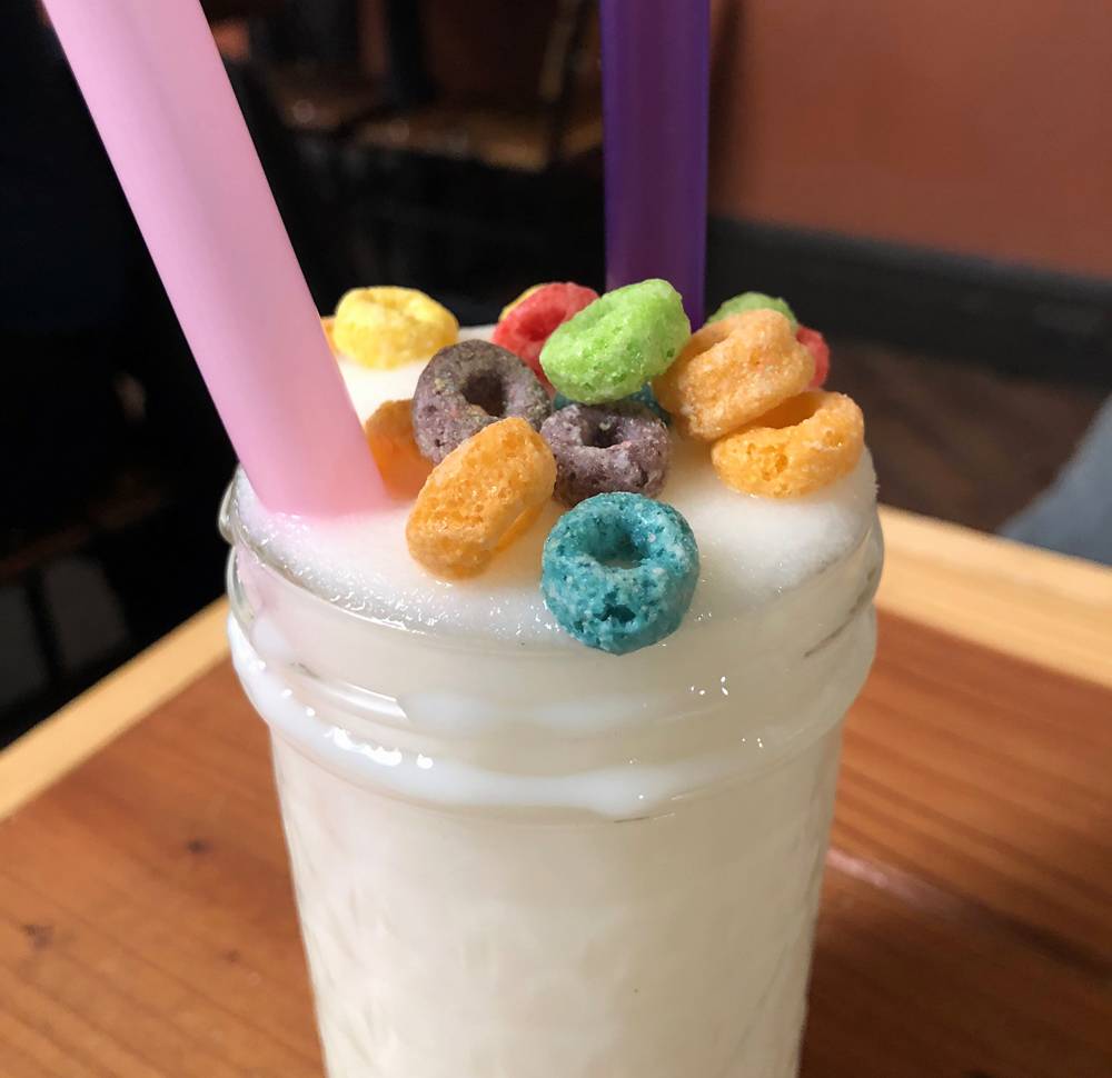 Image: A close up of the top of Watson's Toucan Slam boozy slushie. A frozen, blended white beverage is served in a mason jar. Froot Loopsâ„¢ cereal pieces garnish the drink. A pink straw and a purple straw stick out of the drink on opposite sides of the glass. The glass is on a wood table. Photo by Jessica Hammie.