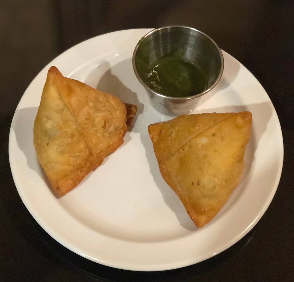 Image: Two pyramid-shaped golden brown samosas sit on a round white plate with a stainless steel condiment cup filled with a cilantro dipping sauce. The plate sits on a black glass table. Photo by Jessica Hammie.