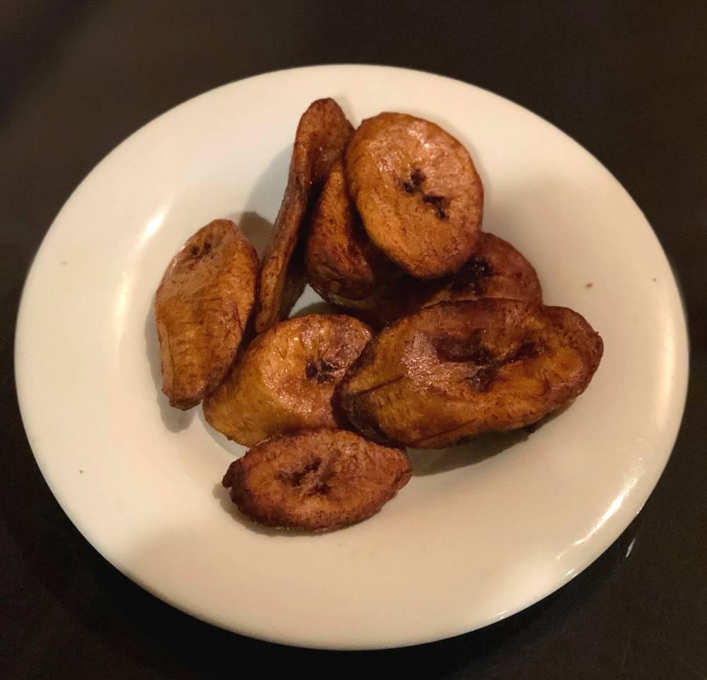Image: Slices of dark golden brown fried plantains sit on a white plate on a black glass table. Photo by Jessica Hammie.