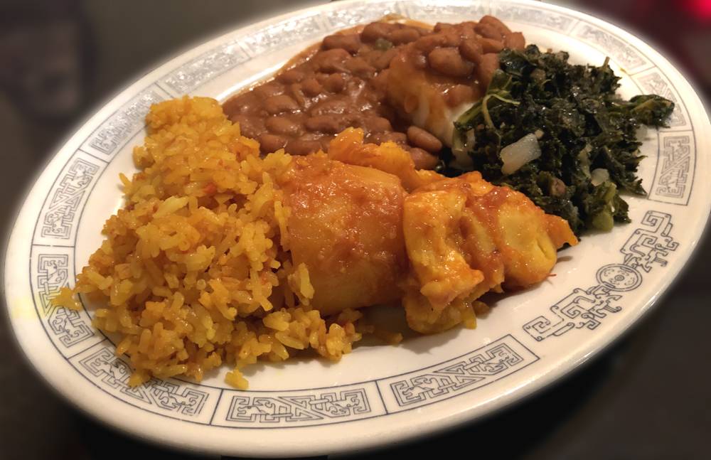 Image: A vegetarian platter includes yellow rice, pinto beans, sautÃ©ed kale, and golden-red sweet potatoes. The plate is an oval shaped and white, with a geometric blue pattern around the edge of the plate. The plate sits on a black glass table. Photo by Jessica Hammie.