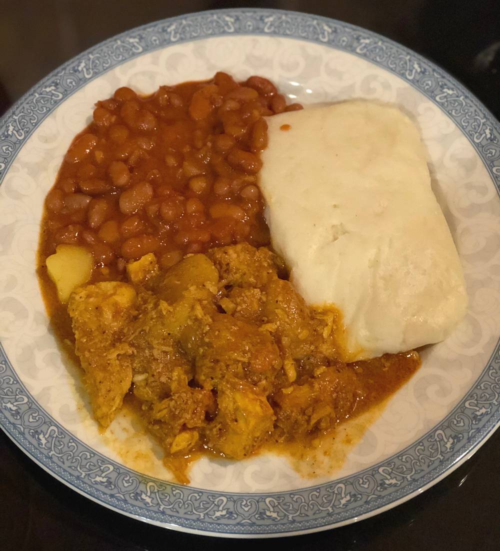 Image: A round white plate with a light blue decorative design on the edge contains pinto beans, nshima, and chicken curry. The plate sits on a black glass table. Photo by Jessica Hammie.