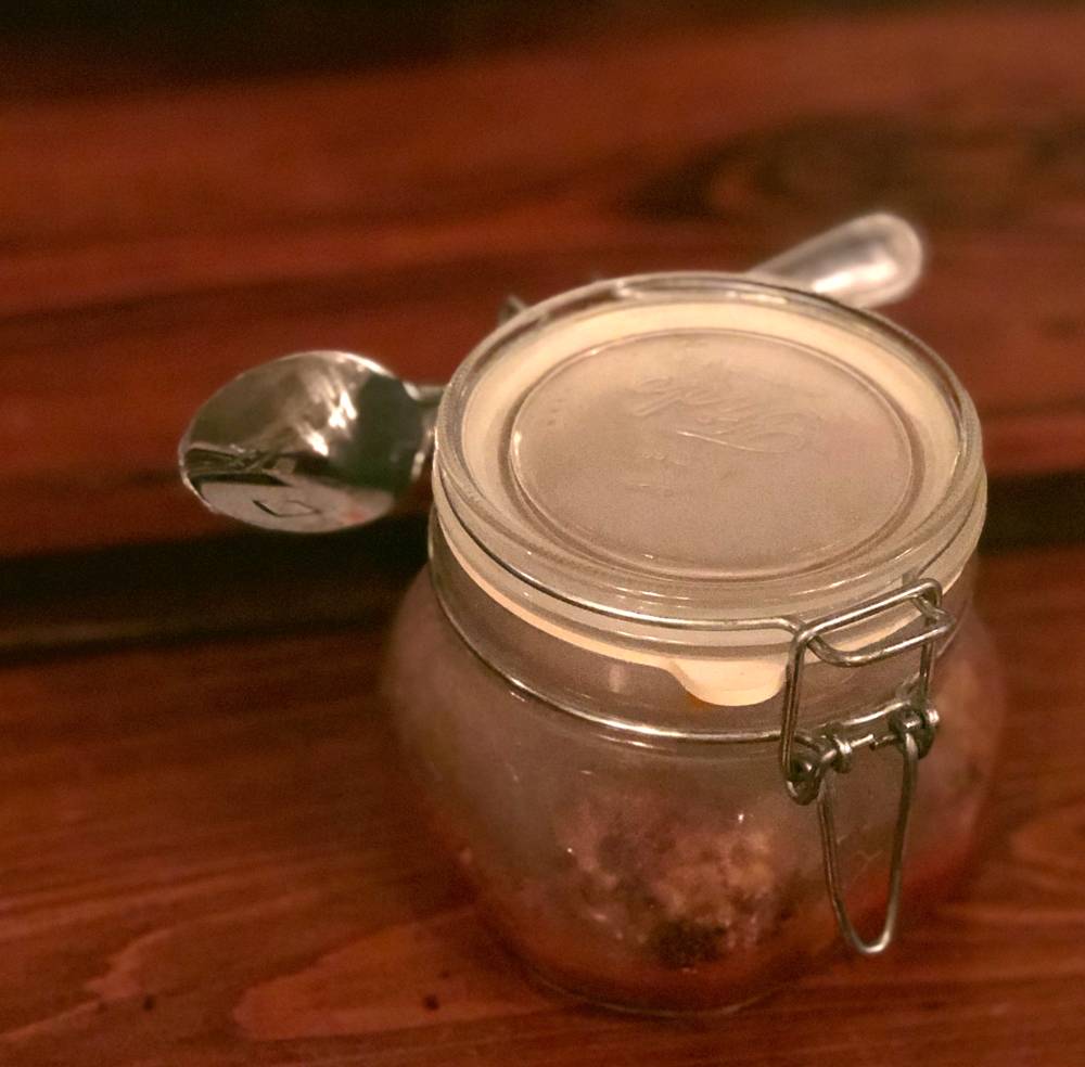 Image: Potato fritter appetizer at the WheelHouse. In a closed, hinged-lid mason jar are potato fritters and marinara sauce. The lid is closed, and the inside of the jar looks smoky. A silver spoon is threaded through the hinge on the back of the jar. The jar sits on a wood table. Photo by Jessica Hammie.