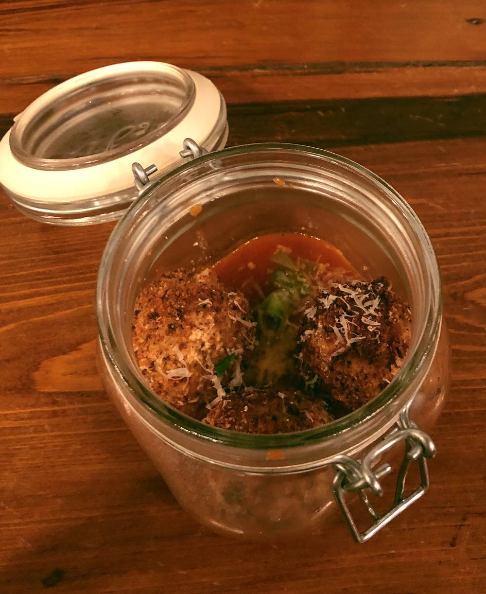 Image: Potato fritter appetizer at the WheelHouse. The hinged-lid mason jar is open. In the jar are three potato fritters in a red marinara sauce. Shaved parmesan dusts the top of the fritters. The jar sits on a wood table. Photo by Jessica Hammie.