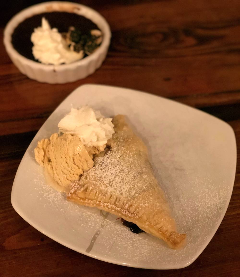 Image: Apple and cherry turnover dessert at the WheelHouse is garnished with cinnamon ice cream and whipped cream, both on the left side of the plate. The turnover is triangularly shaped and sitting on a square white plate on a wood table. Photo by Jessica Hammie.