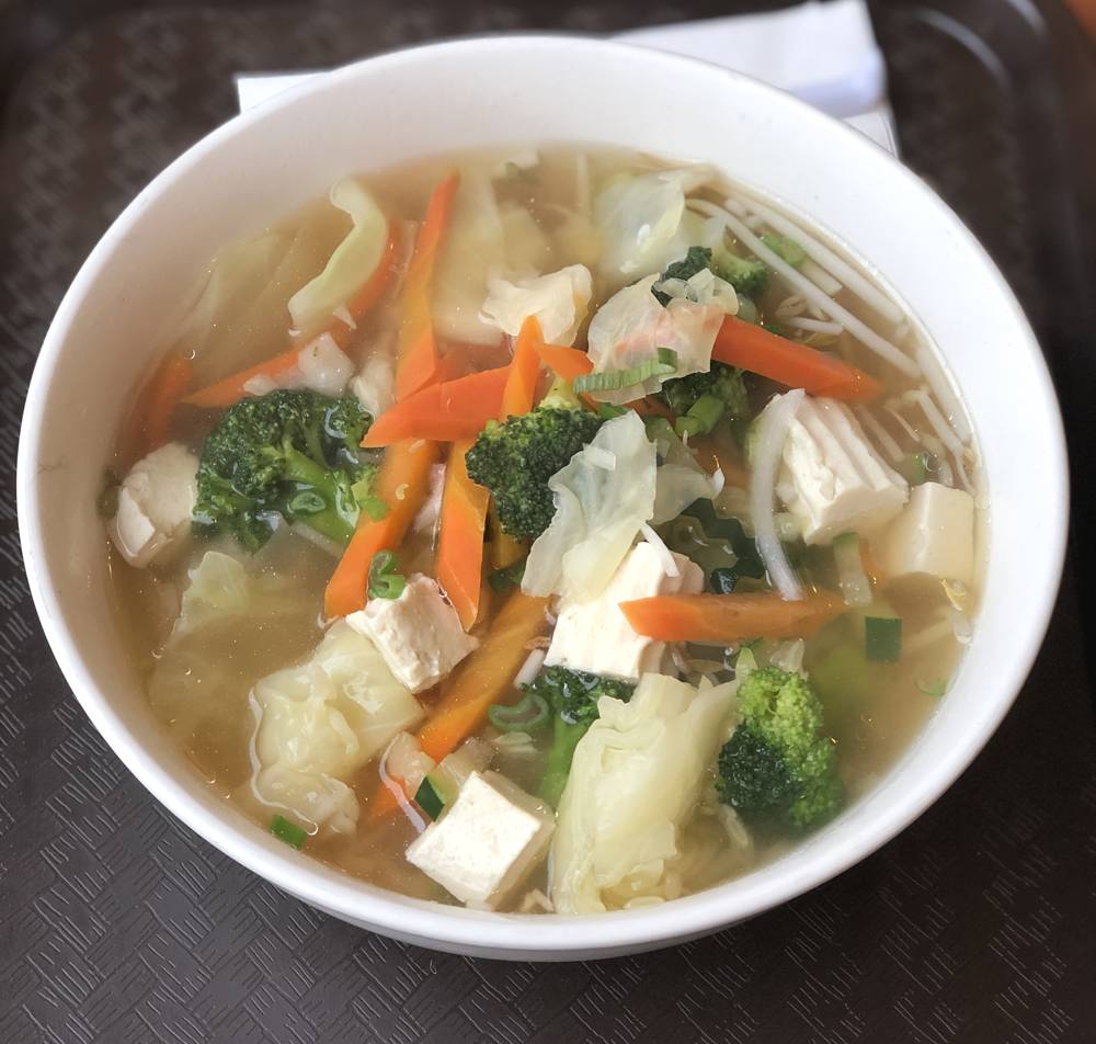 Image: Vegetable noodle soup at Basil Thai. A large white bowl contains tofu, broccoli, carrots, onion, and cabbage in a light colored broth. The bowl is on a dark brown cafeteria tray. Photo by Jessica Hammie. 