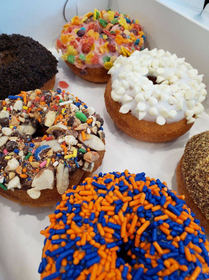 Image: Six donuts are in a white cardboard box, five are mostly visible. They are all cake donuts, and have different toppings: Chocolate, pink icing with Fruity Pebbles, white icing with white chocolate chips, orange and blue sprinkles, crushed candy and nuts. Photo from Facebook event page.