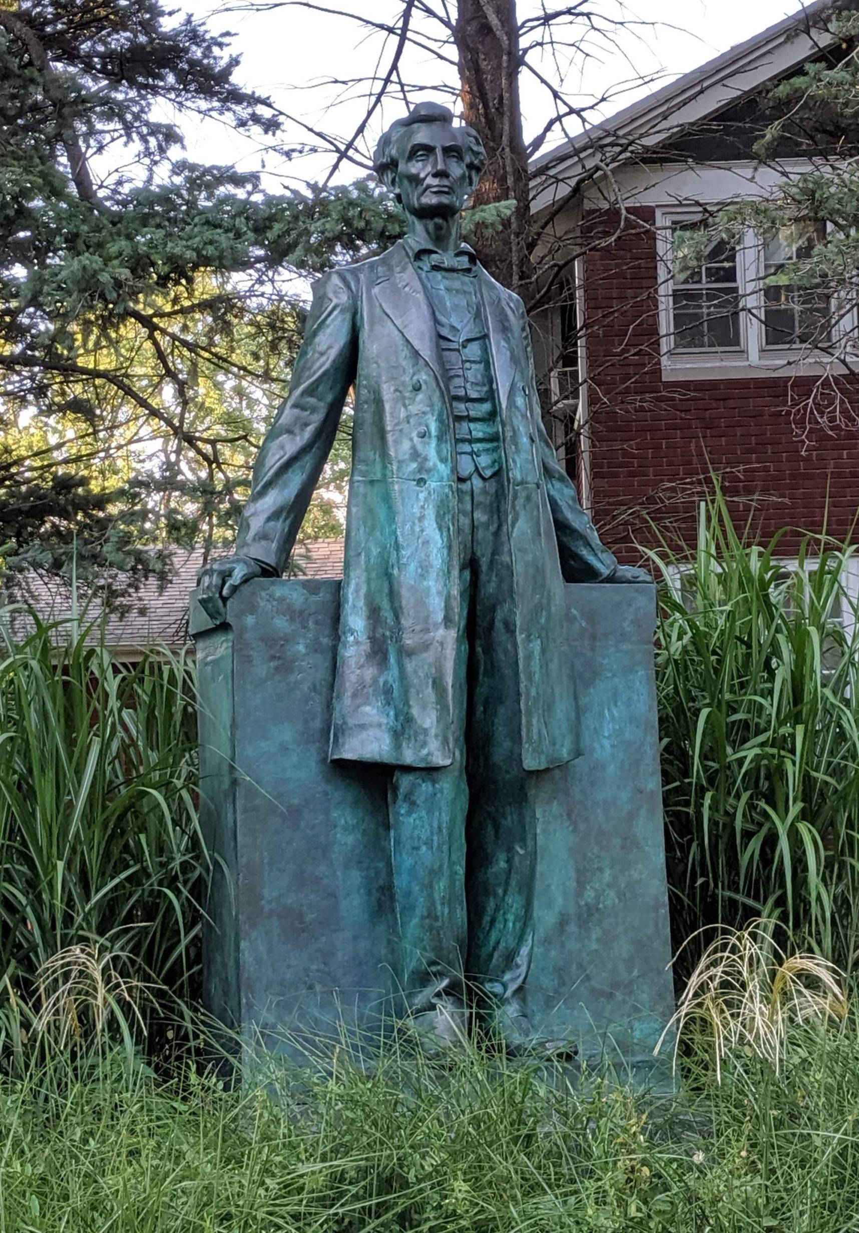 Image: A bronze statue of Abraham Lincoln, now green from oxidation. It is a full length statue, and the figure is leaning on a pedestal. The figure is wearing a long coat and vest. It is surrounded by overgrown grass. A house with red bricks is in the background. Photo by Katriena Knights.