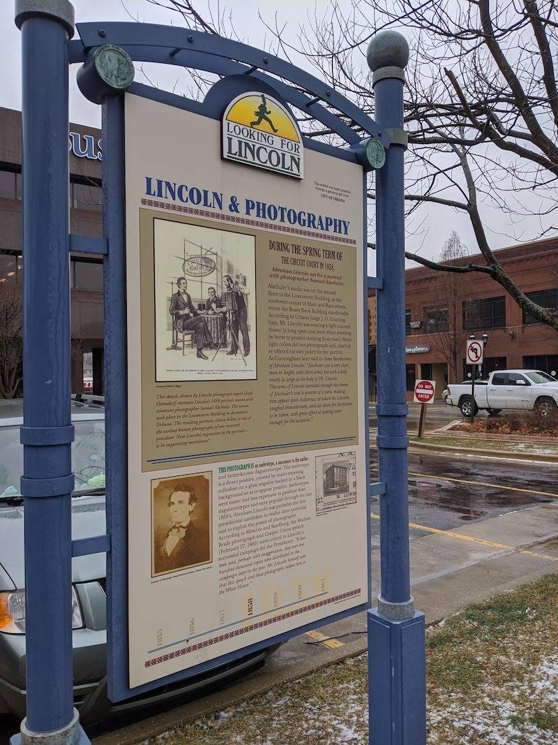 Image: An informational panel with the title Lincoln and Photography. It is framed by blue metal posts. Busey Bank is in the background. Photo by Katriena Knights.