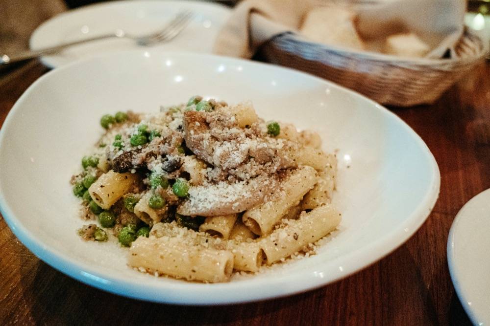 Image: White plate of yellow pasta sits on top of a brown wooden table. There are green peas and grown mushrooms and sausage in the dish. There's a light brown basket in the back right of the photo. Photo by Anna Longworth.