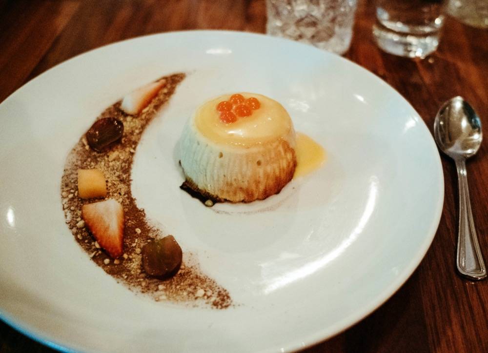 Image: White plate sits on a brown wooden table. Small white dessert sits right of a brown streak of chocolate shaped like a cresent moon. There's a silver spoon right of the plate. Photo by Anna Longworth.