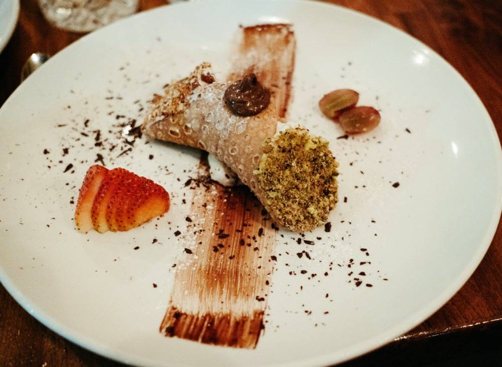 Image: White plate with a brown streak of chocolate down the middle. A brown cannoli sits on top of it, left is a red sliced strawberry. Photo by Anna Longworth.