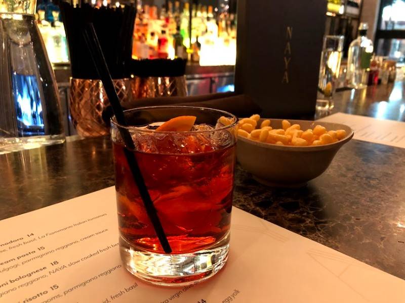 Image: A NAYA Negroni served in a rocks glass is garnished with an orange slice in the beverage and a black cocktail straw. The glass sits atop a menu on a dark marbled bartop. Behind the glass is a bowl of bar snacks and a black cocktail menu that reads 