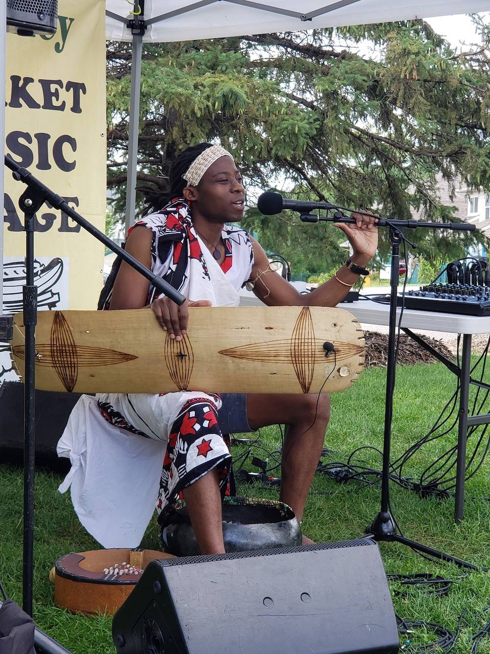 Image: Nyttu Chongo, dressed in shorts and a traditional white wrap with red and black patterns, performs a concert outdoots. He holds a traditional instrument while speaking or singing into a microphone. Sevel instruments sit at his feet. Image from the Facebook event.
