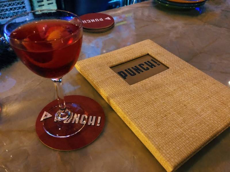 Image: A Punch! Negroni is served in a Nick & Nora glass and garnished with an orange peel in the beverage. The glass sits on a Punch! branded coaster on a gray-brown marbled bartop. To the right of the glass is a menu with 