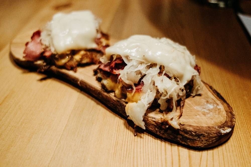 Image: Two sliders of corned beef on top of a live-edge wooden serving plate. The tops of each feature melted white swiss cheese and sauerkraut. The table the dish sits on is a lighter shade of brown wood. Photo by Anna Longworth.