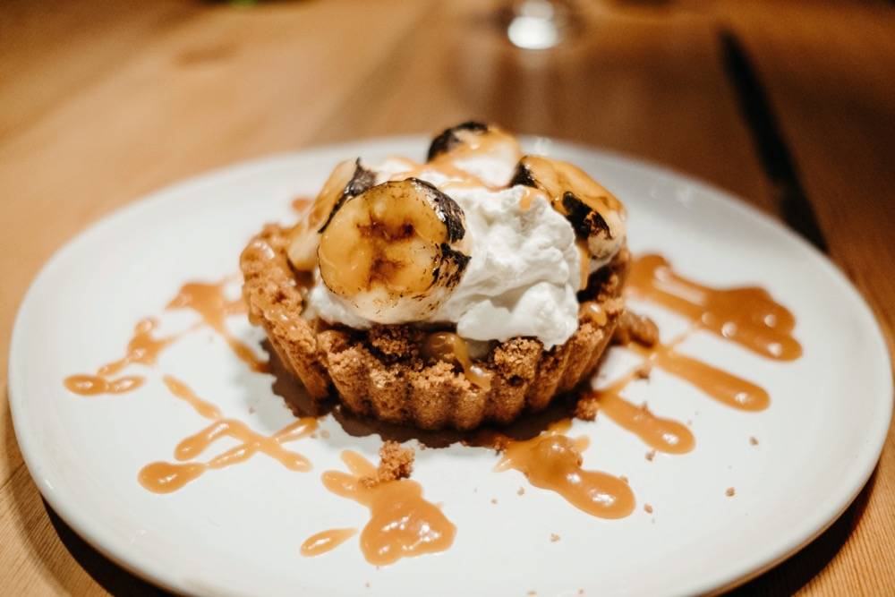Image: White plate with a small brown pie crust with white whipped cream and bananas. There is brown sauce drizzled across the plate. Photo by Anna Longworth.