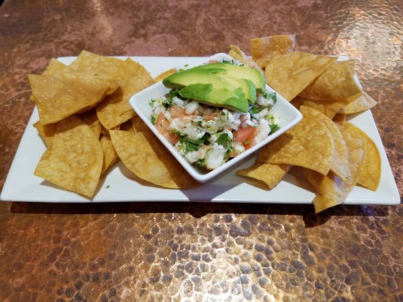 Image: Shrimp ceviche is garnished with avocado slices and served in a small, square, white bowl. The bowl is in the middle of a rectagular white plate that holds golden tortilla chips. The plate sits on a hammered copper table.  Photo by Matthew Macomber. 