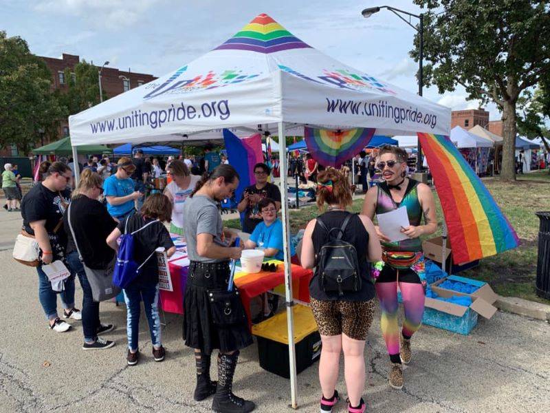 Image: A white tailgate tent with rainbow stripes at the top. It says www.unitingpride.org on the two visible edges. A rainbow flag is hanging from the tent, and there is a blue, purple, and pink striped flag on the opposite side. Several people are gathered around a table. Photo from Uniting Pride Facebook page. 