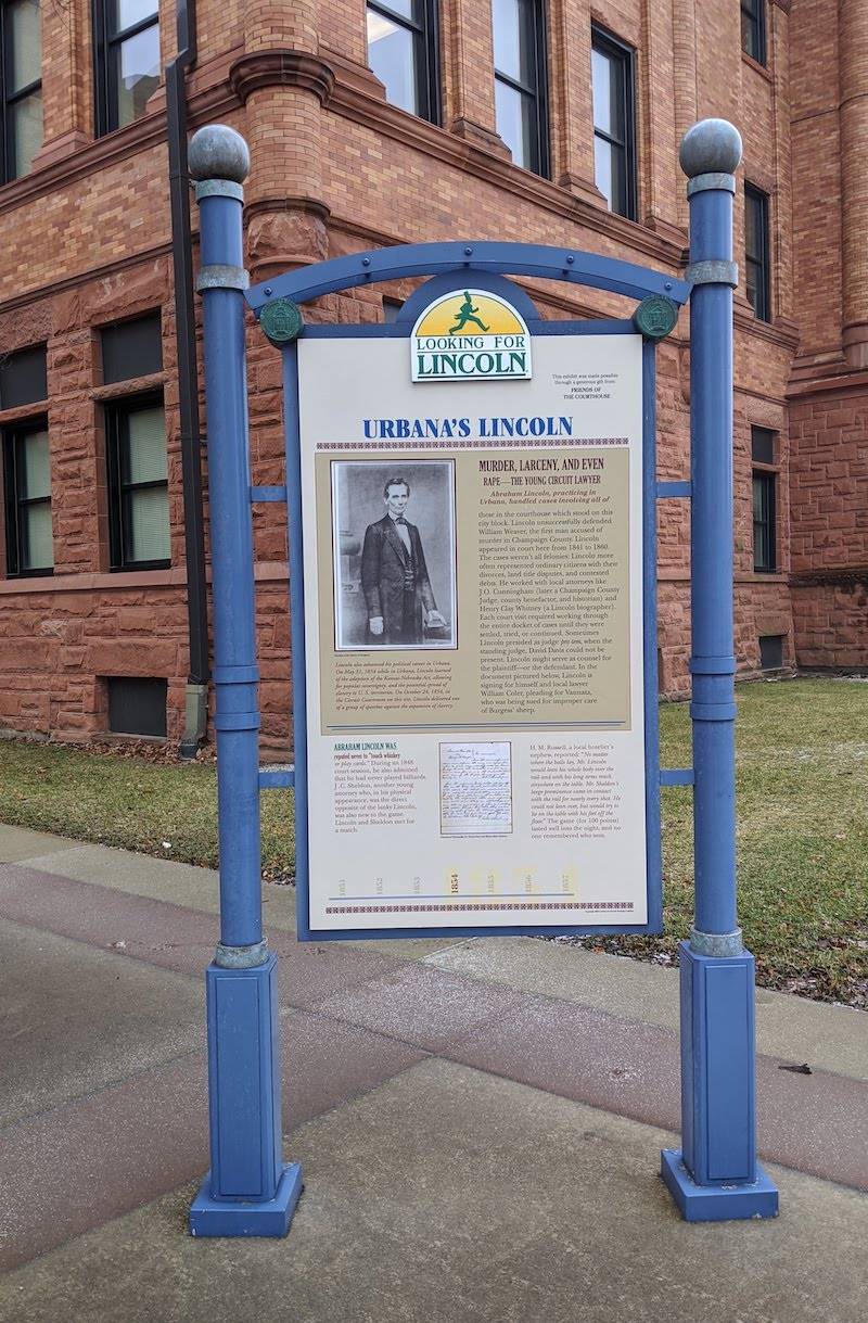Image: A close up of an informational panel with the title Urbana's Lincoln. It's framed by blue posts. The courthouse is in the background. Photo by Katriena Knights. 