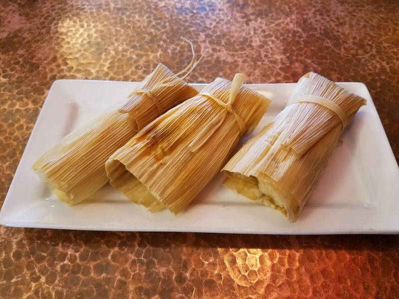 Image: Three corn-husk wrapped tamales sit on a rectangular white plate. The tamales are a light golden brown, and rectangular. The plate sits on a hammered copper table. Photo by Matthew Macomber. 