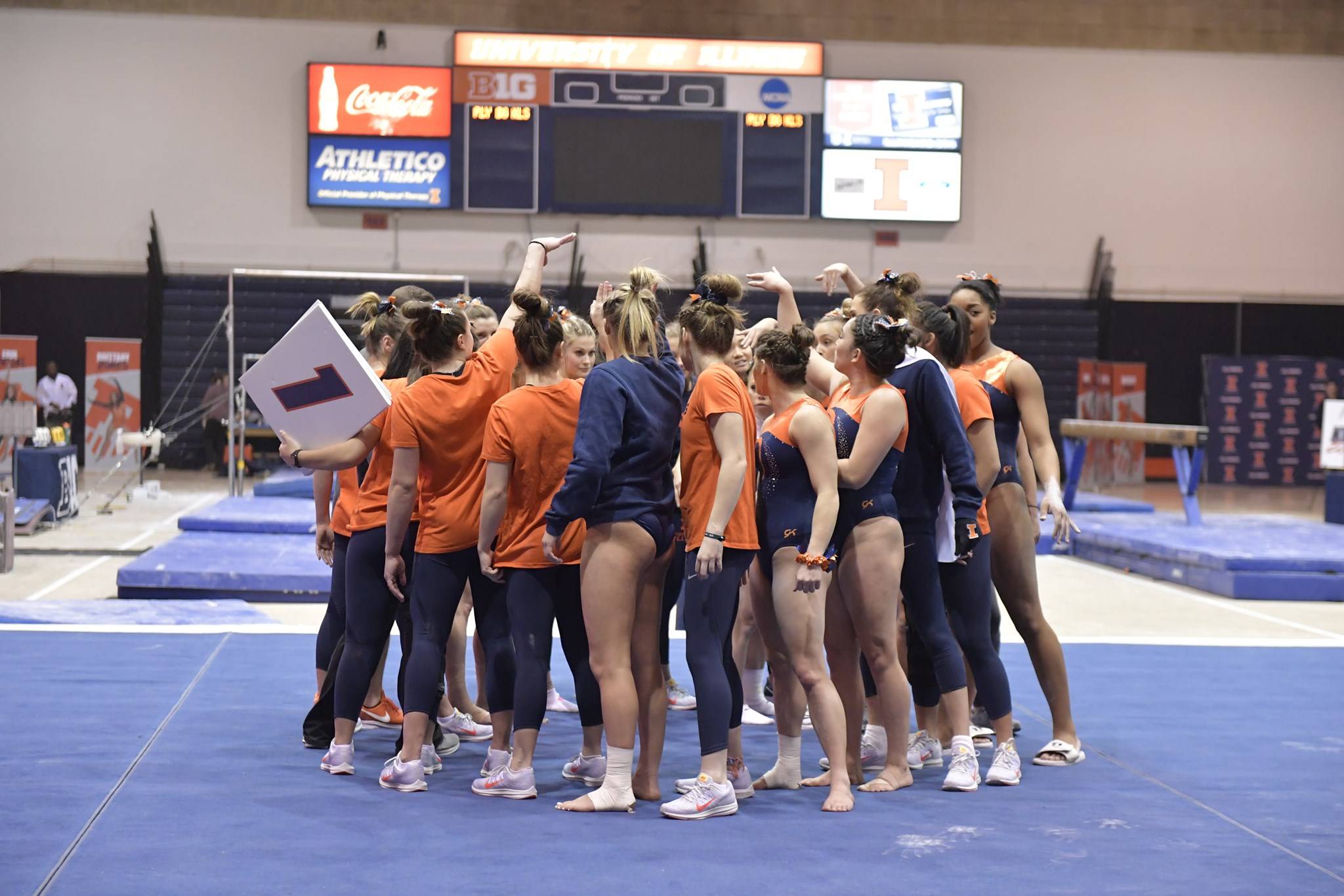 A huddle of the University of Illinois womenâ€™s gymnastics team standing on a blue mat with their backs to the camera. A few of them have their arms in the air. They are standing in a gym space, and a score board is visible on the wall in the back. Photo from the Illini womenâ€™s gymnastics vs. Central Michigan Facebook event page.