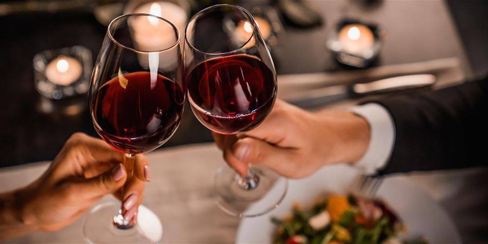 Image: Stock image of a manâ€™s hand and a womanâ€™s hand clinking wine glasses filled with red wine. The background is blurry, but a dinner table with candles and a dish of food can be seen. Photo from the Facebook event page.