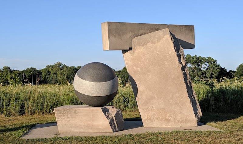 A stone sculpture with two separate parts. There is a thick stone slab with a dark gray sphere sitting on top. Next to it is a tall vertical rectangular stone tilted towards the other structure. It has a horizontal rectangle resting in a notch at the top. Photo by Katriena Knights. 