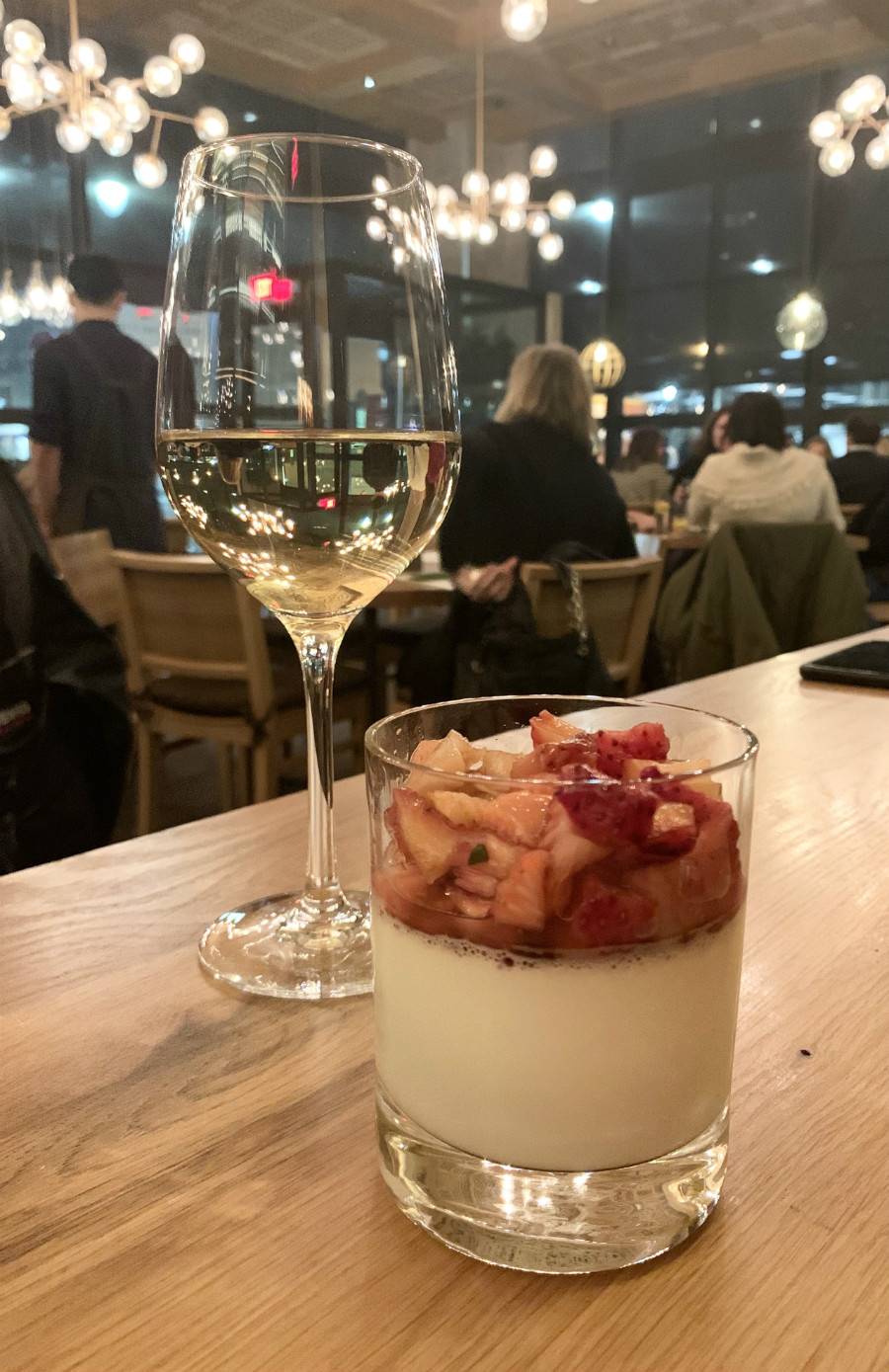 A tumbler glass is half filled with vanilla panna cotta at NAYA. Mascerated strawberries are piled on top and near the top of the glass. Behind the panna cotta is a glass of white wine. Both glasses sit on a light colored wood table. Photo by Stephanie Wheatley. 