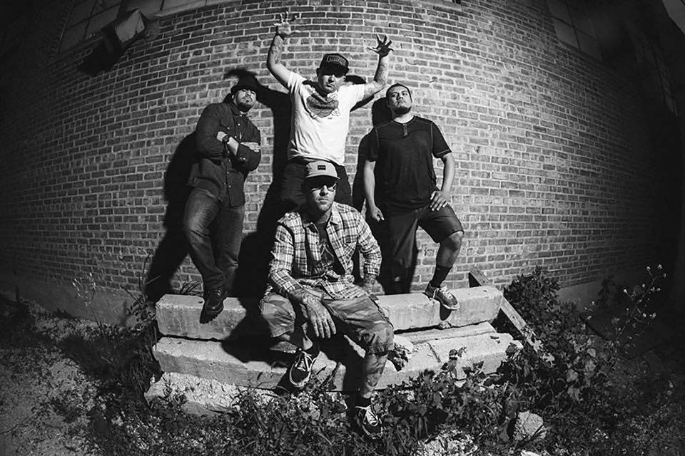 Black and white photograph taken with a fish-eye lens. Four men sit and stand on worn down concrete stairs against a brick wall. Three men stand in the back, with one man crossing his arms, the one in the middle holding his arms above his head, fingers out stretched like claws, and a third stands with one hand on his front leg. In the foreground, a man sit on the stairs looking into the camera.