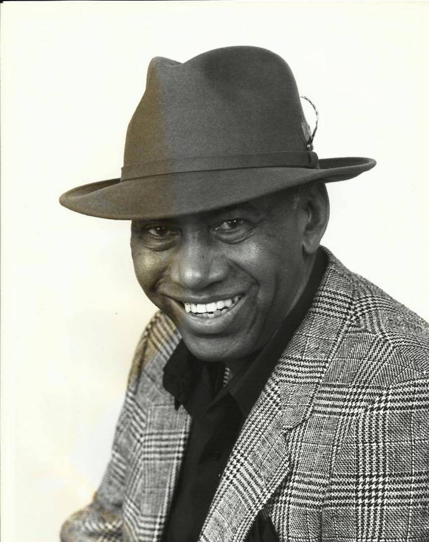 Image: Candy Foster sits, smiling to the camera. He wears a traditional fedora hat with a feather, a black shirt, and a herringbone suit coat. The image is in sepia tones. Image from the Facebook event.