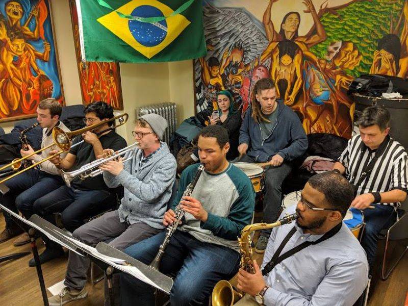 Two rows of people playing a variety of instruments: saxophone, trombone, trumpet, clarinet, drums. There is a Brazilian flag hanging from the ceiling, and colorful murals on the walls behind them. Photo from Luso-Brazilian Student Assocation Facebook page.