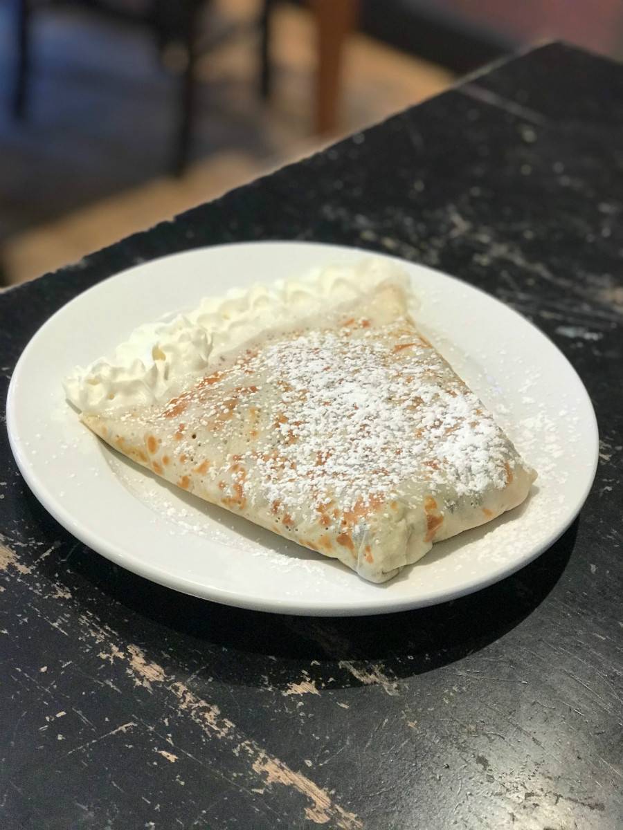 At Pekara, a folded crepe is served on a round, white plate with whipped cream along the open edge of the crepe. The crepe has golden brown spots and is dusted with powdered sugar. The plate sits on a worn, painted black table. Photo by Stephanie Wheatley. 