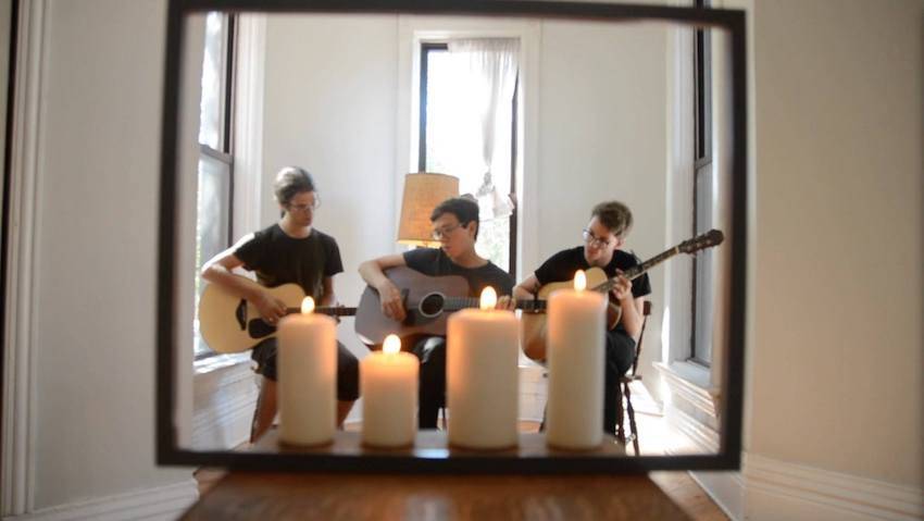 The reflection of three guitar players sitting together with their instruments in a well-lit room with tall windows, white curtains, and white walls. Four candles are also reflected in the mirrored image in front of the guitarists.